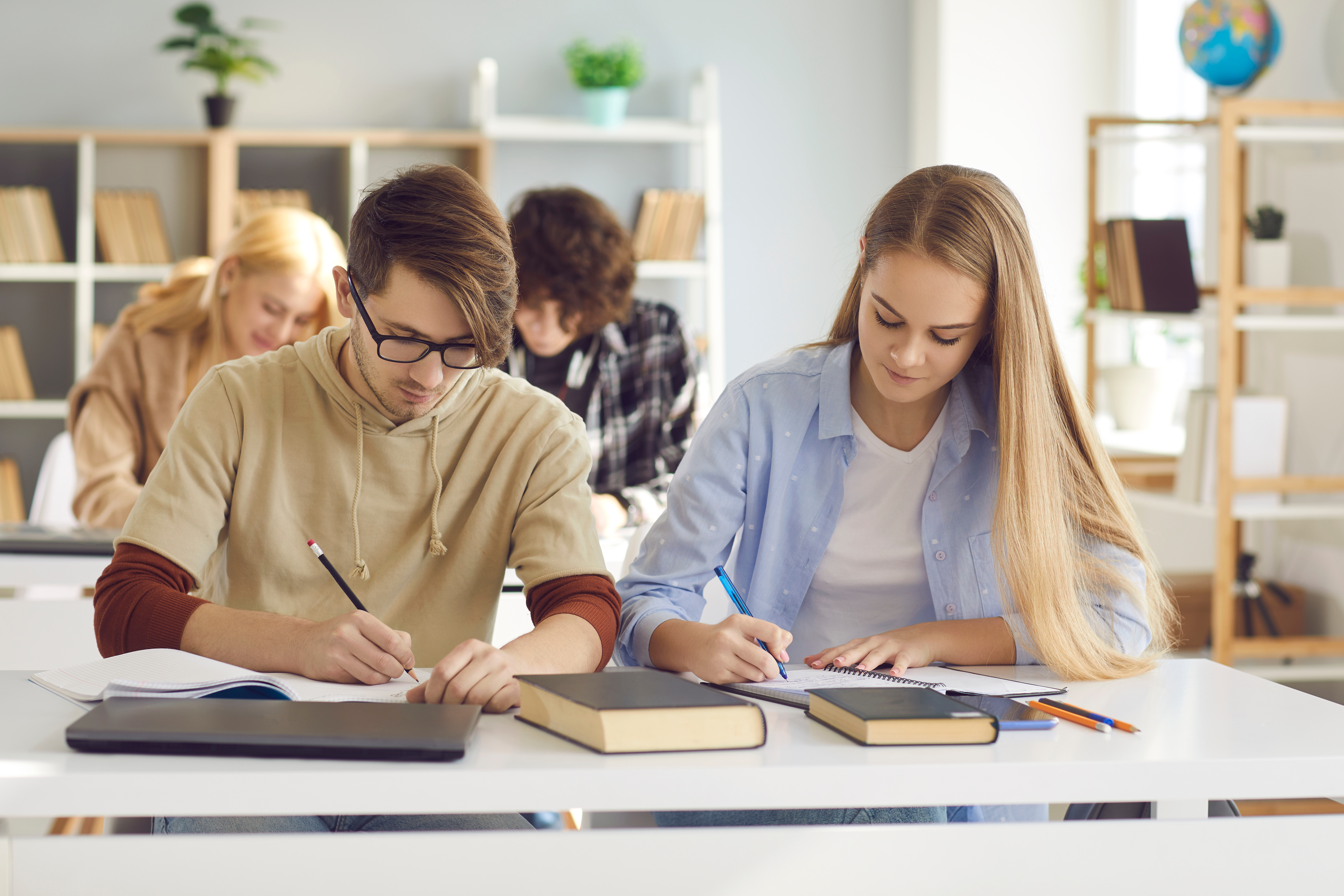 Studenti sjede u učionici. Vidimo studenta i studenticu u klupi kako pišu u bilježnice. Ispred njih su dvije knjige i laptop.