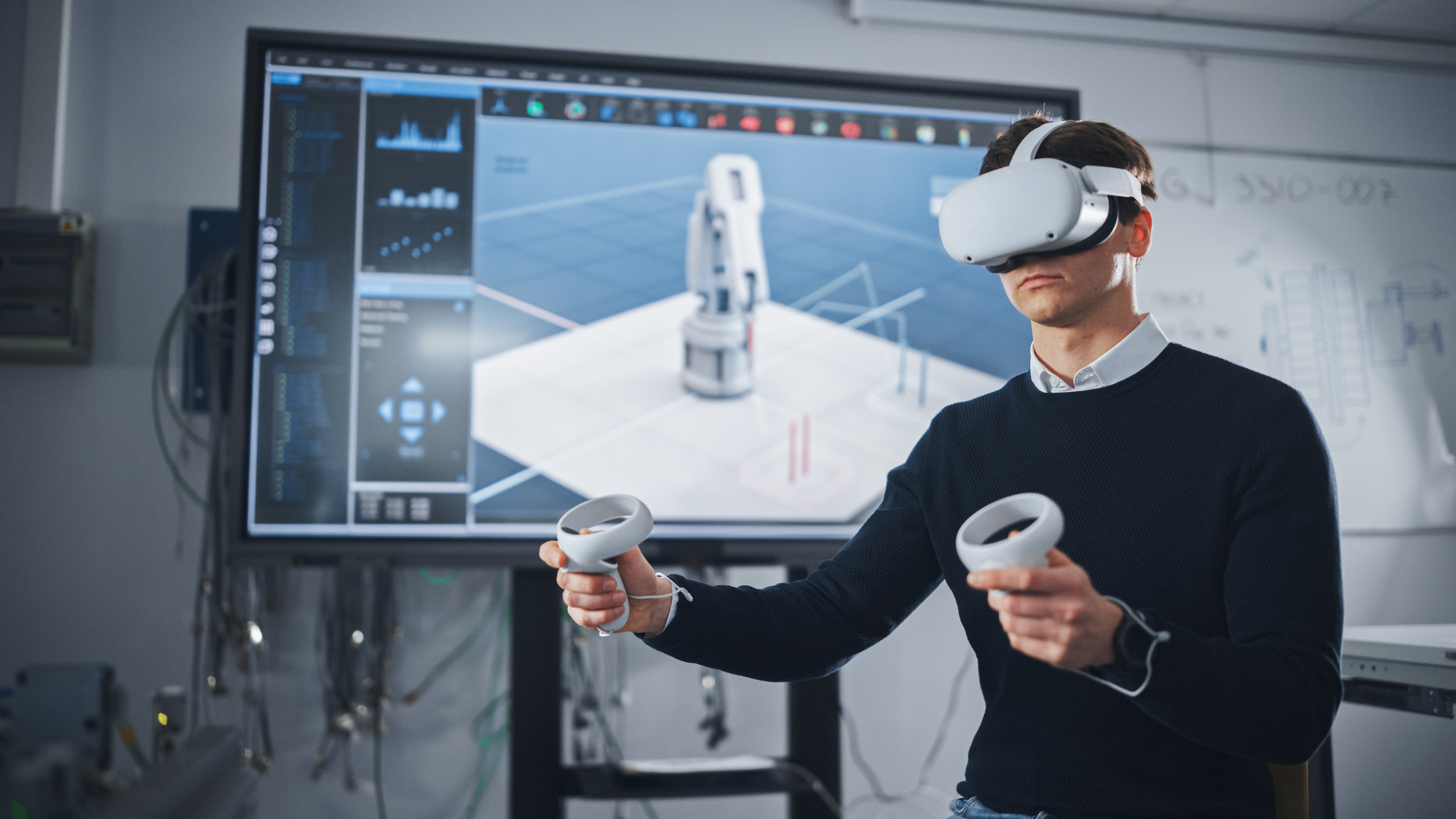 Mladi inženjer nosi VR naočale i drži kontrolere ispred ekrana s 3D modelom robotske ruke