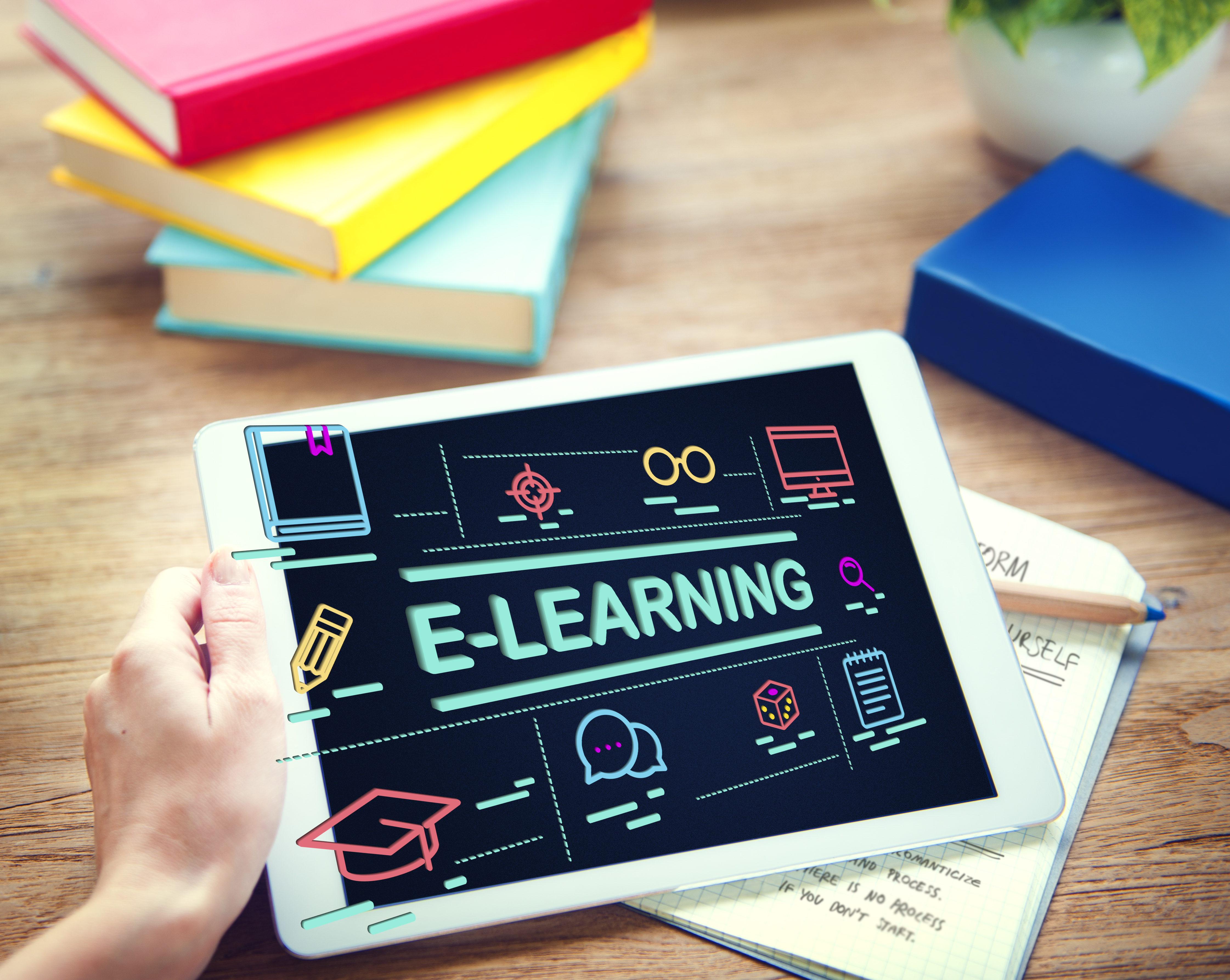 Ruka drži tablet s natpisom E-LEARNING i ikonama učenja, okružena šarenim knjigama i bilježnicom na stolu