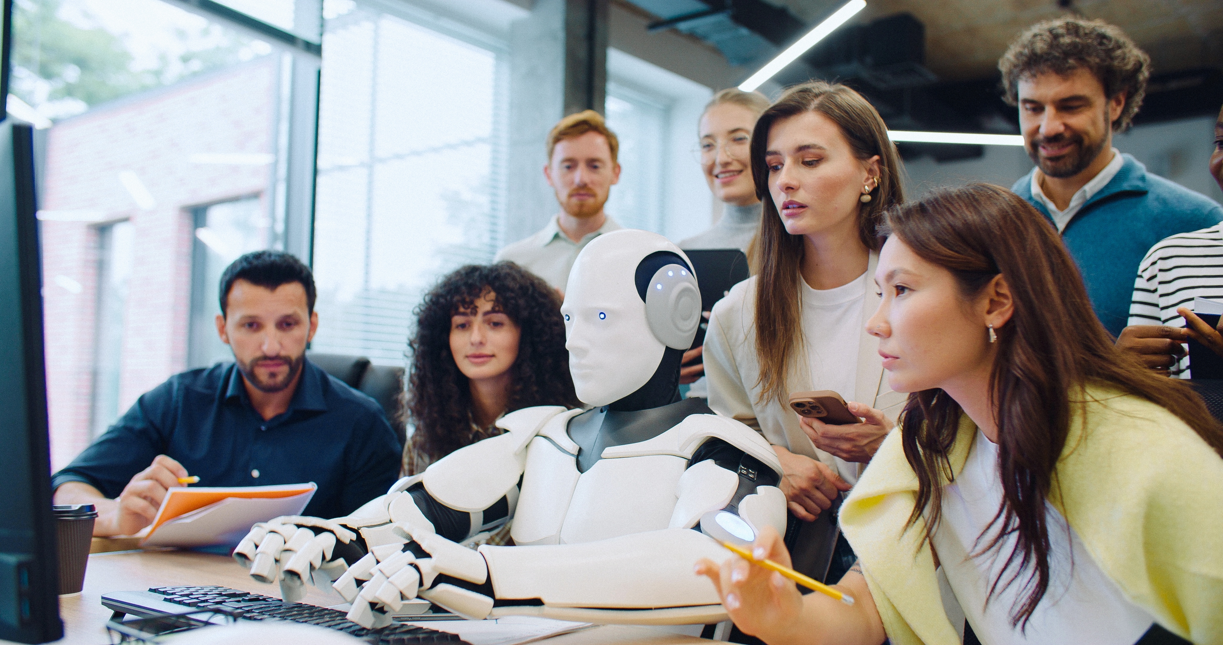 Grupa zaposlenika promatra robota humanoida koji radi na računalu za stolom u modernom uredu