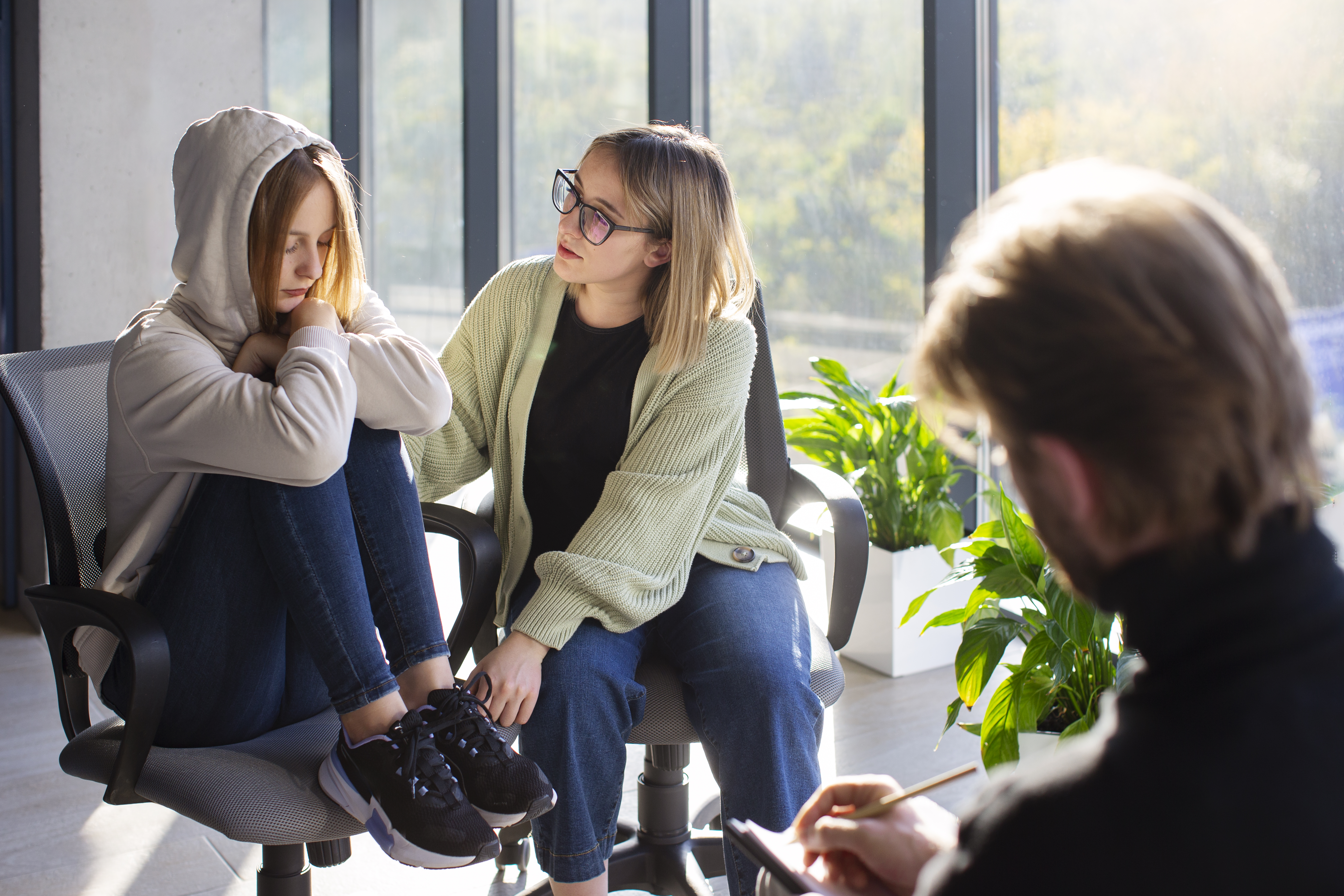 A female student sitting on a chair with her legs tucked in and a hood on her head, looking worried, being comforted by another student