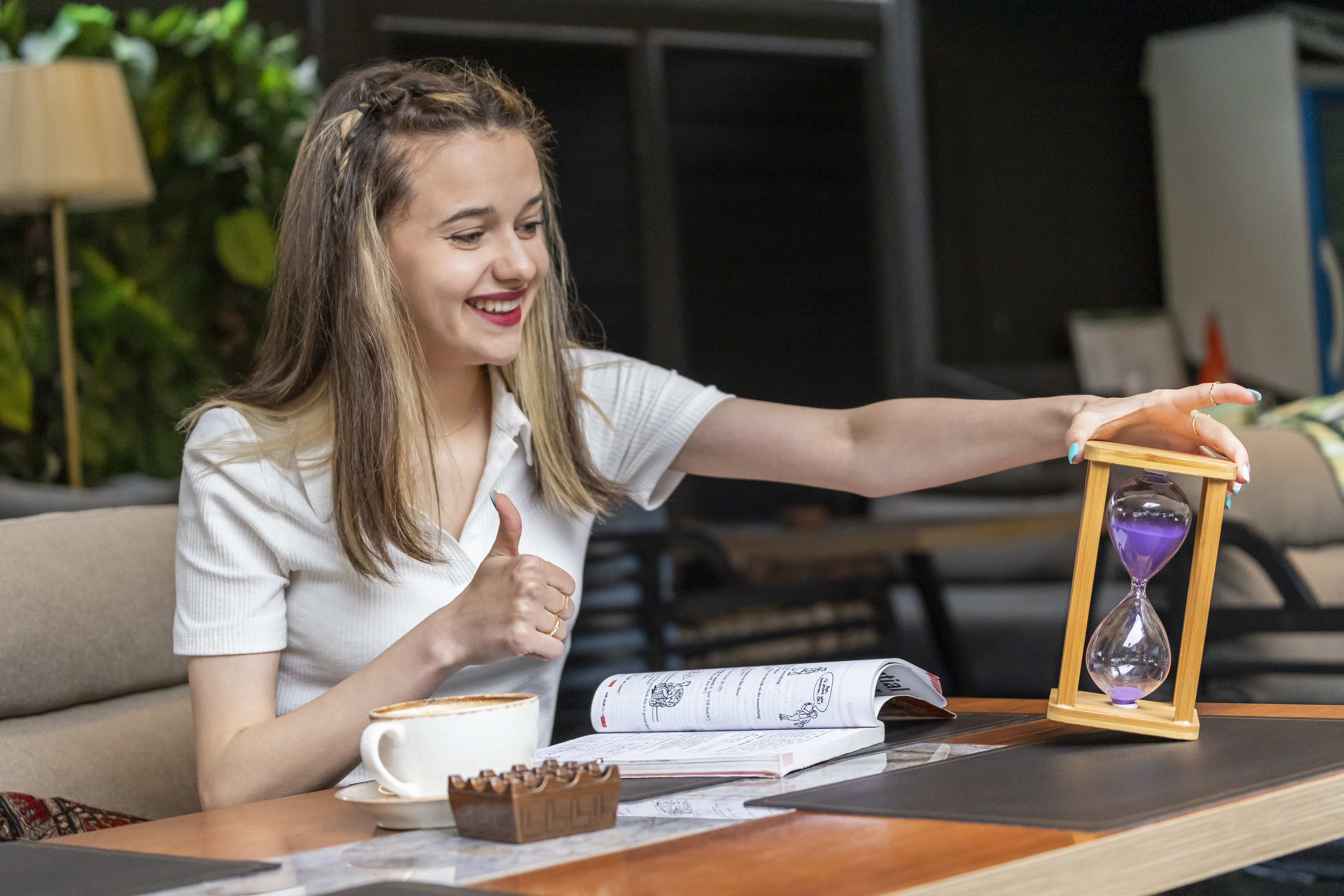 A girl sitting at a table with coffee and notebooks, holding an hourglass and showing a thumbs up